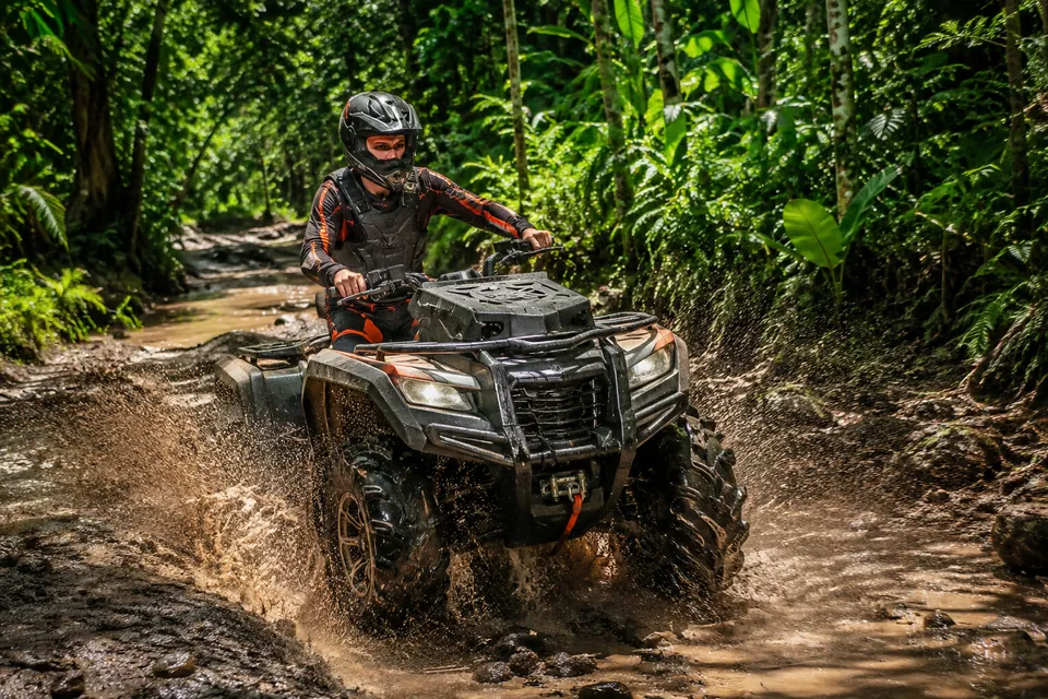 A helmeted rider steering an ATV along a muddy jungle trail