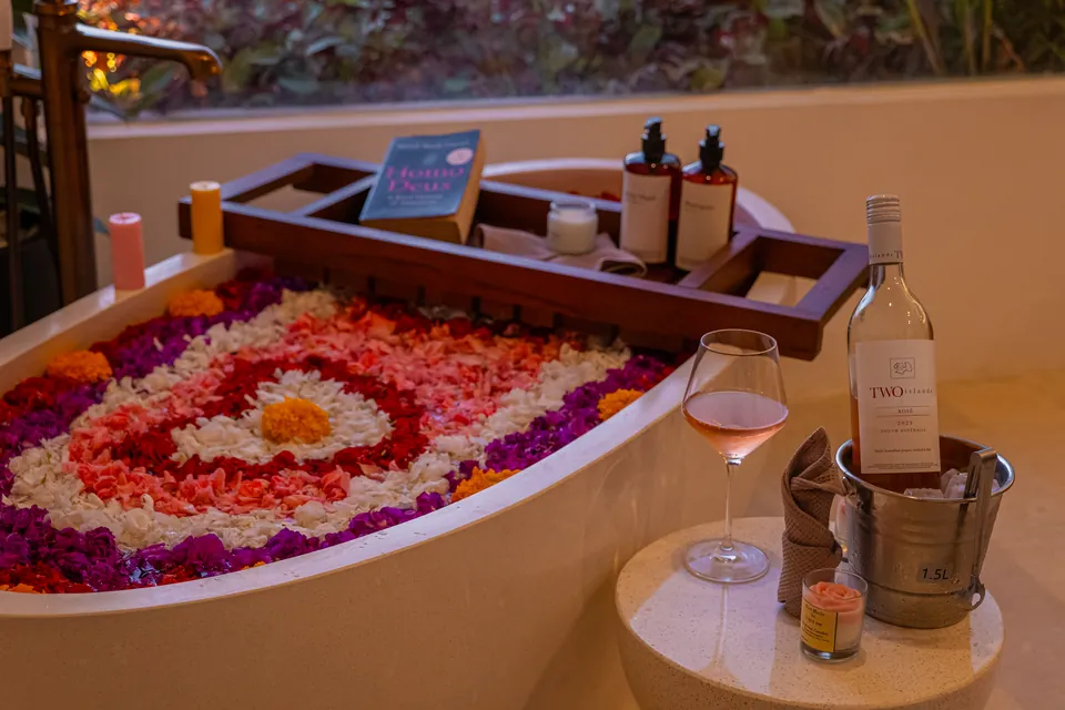 A petal-filled bathtub at dusk, with a bath tray, candles and a bottle of rosé on ice