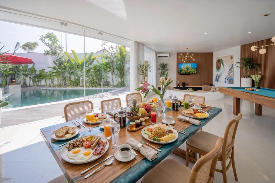 A long villa table by the glass doors to the pool, set with breakfast for several guests: eggs, toast, fried rice, fresh fruit, juices and coffee
