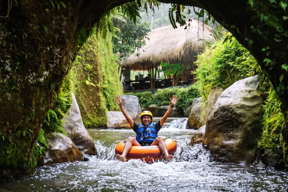A smiling guest in a helmet and life jacket floating on an inner tube under a rock arch